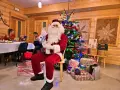 Święty Mikołaj wita gości podczas pobytu z kolacją wigilijną. W tle choinka i prezenty.
