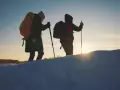 Dwie osoby z plecakami i kijkami trekkingowymi idą przez ośnieżony teren. W tle zachodzące słońce, przypominające czerwone wierchy szczyty.