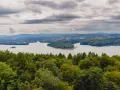 Widok na jezioro otoczone lasami, z górami w tle. Na pierwszym planie bujna zieleń drzew, a dalej spokojna tafla wody i zabudowania. Nad wszystkim chmury, jakby biała góra na niebie.