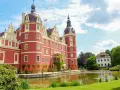 Piękny czerwony zamek otoczony wodą i zielenią. Idealne miejsce na zwiedzanie parku mużakowskiego.