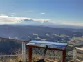 Widok na pasmo górskie z tablicą informacyjną i kijkami trekkingowymi. W oddali majaczą ośnieżone szczyty, a poniżej rozciąga się malowniczy krajobraz z lasami i polami, przypominający piękno **wąwozu Homole**.