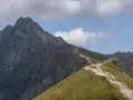Kamienista ścieżka wije się pośród zielonych zboczy, prowadząc do skalistych szczytów w Tatrach. W oddali widać majestatyczne góry, a na szlaku spacerują turyści.