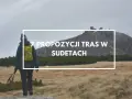 Turystka z plecakiem i kijkami maszeruje przez trawiaste tereny w Sudetach, zmierzając ku górze z budynkami.