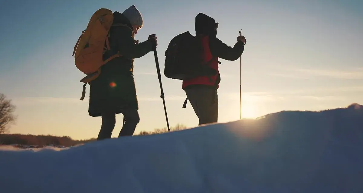 Dwie osoby z plecakami i kijkami trekkingowymi idą przez ośnieżony teren. W tle zachodzące słońce, przypominające czerwone wierchy szczyty.