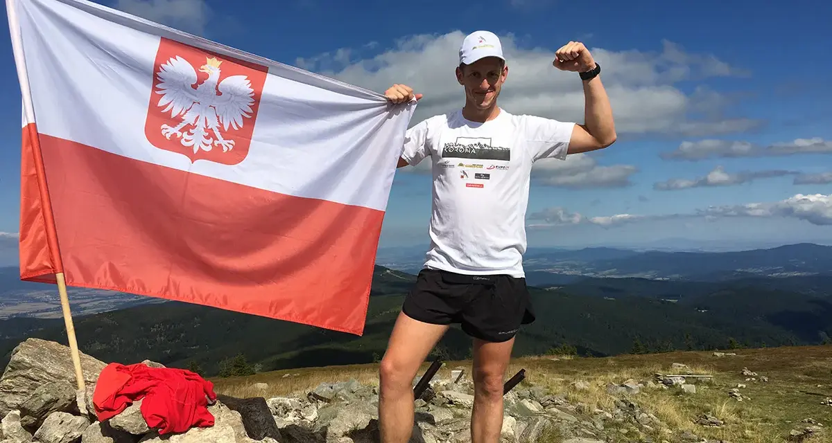 Mężczyzna z polską flagą na szczycie górskim, zdobywca diadem polskich gór, dumny z osiągnięcia.