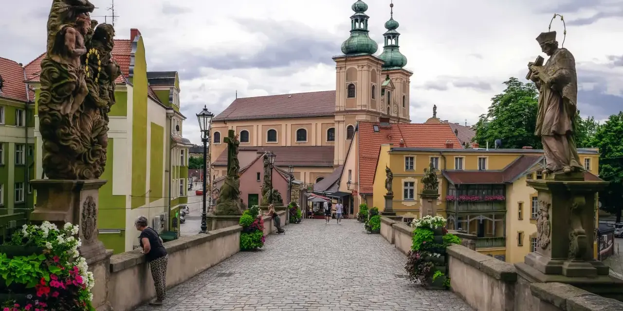 Kamienna, brukowana ścieżka prowadzi do barokowego kościoła z zielonymi wieżami. Wzdłuż mostu stoją kamienne rzeźby. To piękne ziemia kłodzka atrakcje.