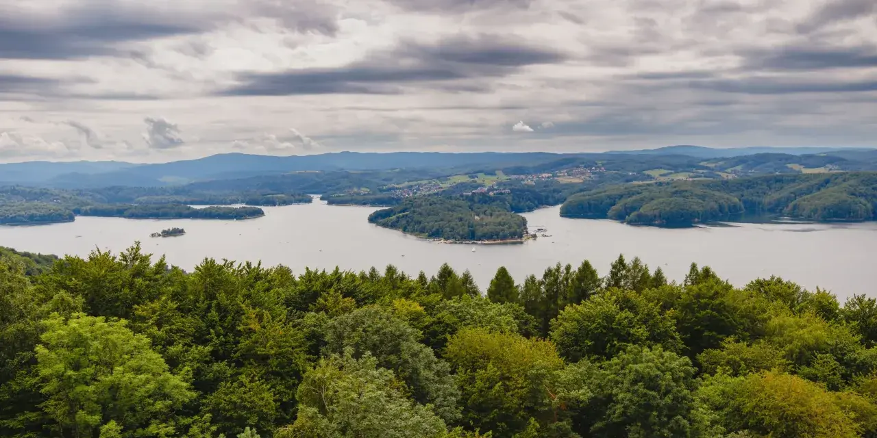 Widok na jezioro otoczone lasami, z górami w tle. Na pierwszym planie bujna zieleń drzew, a dalej spokojna tafla wody i zabudowania. Nad wszystkim chmury, jakby biała góra na niebie.