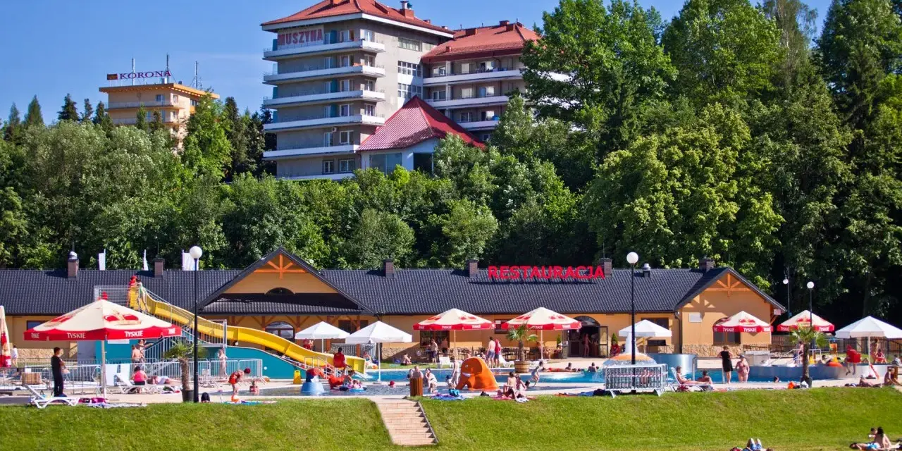 Słoneczny dzień w Muszynie-Zdroju. Ludzie relaksują się przy basenie zjeżdżalnią, a w tle widać hotel i restaurację.