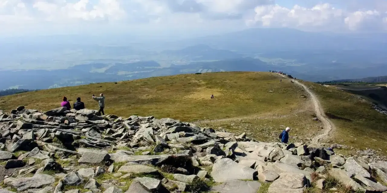 Kamienista ścieżka na szczycie, z której roztacza się widok na doliny. Turyści podziwiają krajobraz, zastanawiając się, ile ma Babia Góra.