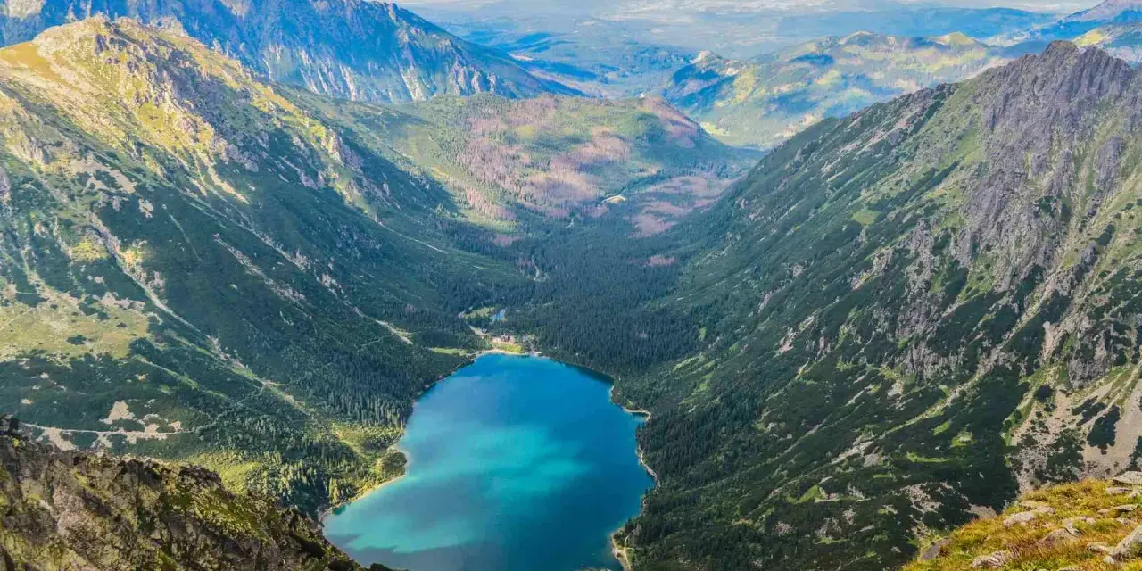 Widok na Morskie Oko z góry. W dole błękitna tafla jeziora otoczona lasem i górami. Kamera uchwyciła parking w oddali.