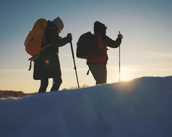 Dwie osoby z plecakami i kijkami trekkingowymi idą przez ośnieżony teren. W tle zachodzące słońce, przypominające czerwone wierchy szczyty.