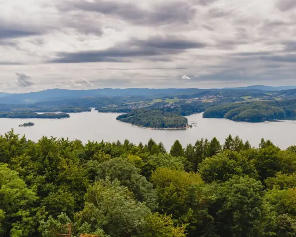 Widok na jezioro otoczone lasami, z górami w tle. Na pierwszym planie bujna zieleń drzew, a dalej spokojna tafla wody i zabudowania. Nad wszystkim chmury, jakby biała góra na niebie.