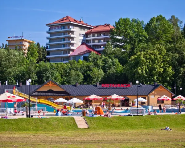 Słoneczny dzień w Muszynie-Zdroju. Ludzie relaksują się przy basenie zjeżdżalnią, a w tle widać hotel i restaurację.
