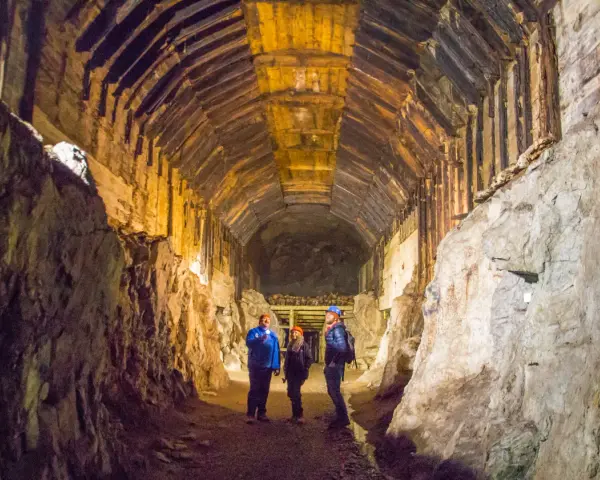 Zwiedzanie podziemnego kompleksu Projekt Riese. Trzech turystów w kaskach oświetla latarkami mroczny tunel z drewnianymi stropami.