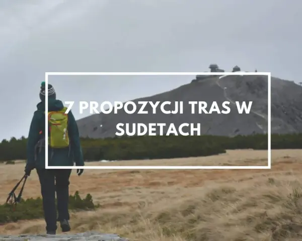 Turystka z plecakiem i kijkami maszeruje przez trawiaste tereny w Sudetach, zmierzając ku górze z budynkami.