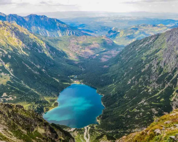 Widok na Morskie Oko z góry. W dole błękitna tafla jeziora otoczona lasem i górami. Kamera uchwyciła parking w oddali.