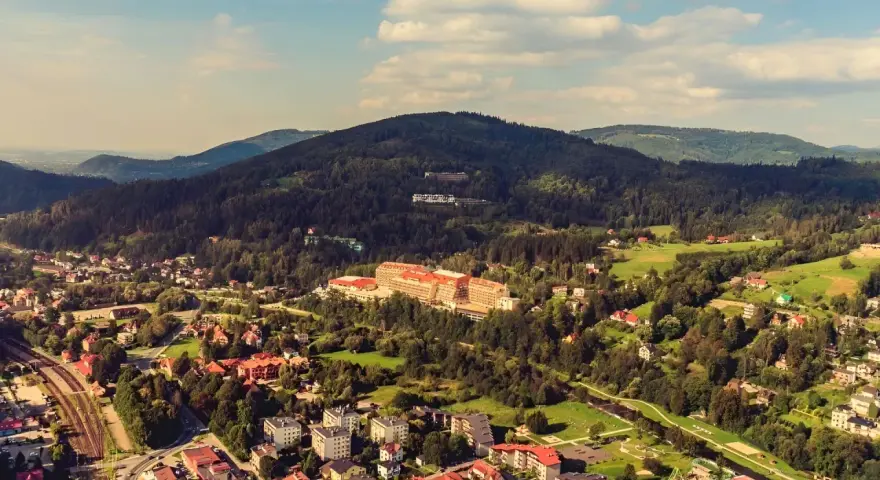 Widok z kamery na hotel Gołębiewski w Wiśle, otoczony zielonymi wzgórzami i zabudowaniami.