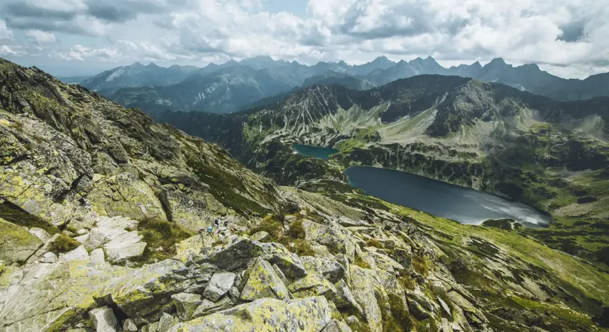 Kamienista ścieżka w górach z widokiem na dwa jeziora i pasmo górskie w tle.