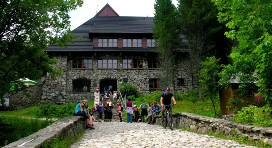 Kamienny budynek schroniska w Dolinie Chochołowskiej z ludźmi na schodach i rowerzystą na kamiennej drodze.