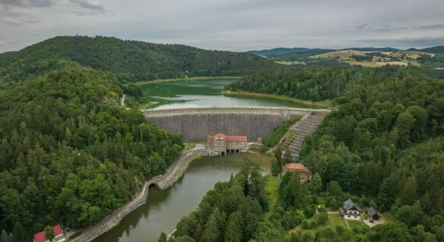 Widok z lotu ptaka na zalesione wzgórza i dużą kamienną zaporę wodną z budynkami.