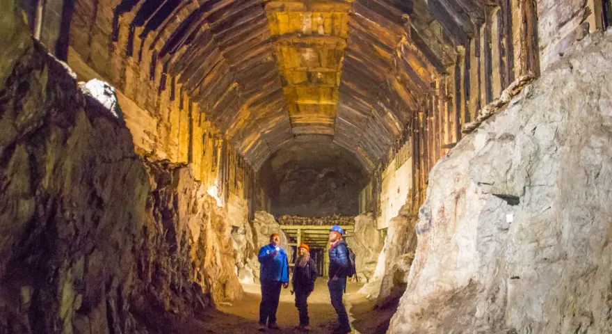 Zwiedzanie podziemnego kompleksu Projekt Riese. Trzech turystów w kaskach oświetla latarkami mroczny tunel z drewnianymi stropami.