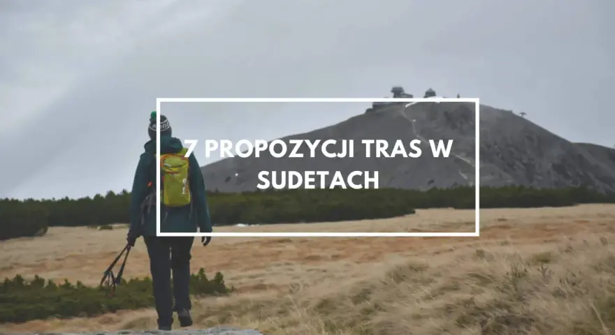 Turystka z plecakiem i kijkami maszeruje przez trawiaste tereny w Sudetach, zmierzając ku górze z budynkami.
