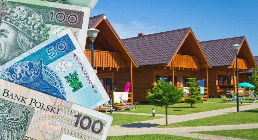 Polskie banknoty przed drewnianymi domkami letniskowymi. Zastanawiasz się, gdzie szukać hoteli? Tu możesz znaleźć nocleg.