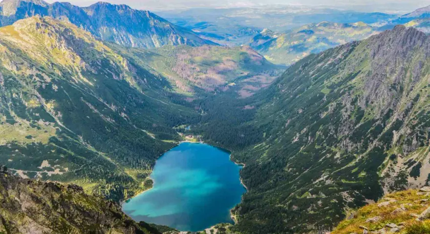 Widok na Morskie Oko z góry. W dole błękitna tafla jeziora otoczona lasem i górami. Kamera uchwyciła parking w oddali.