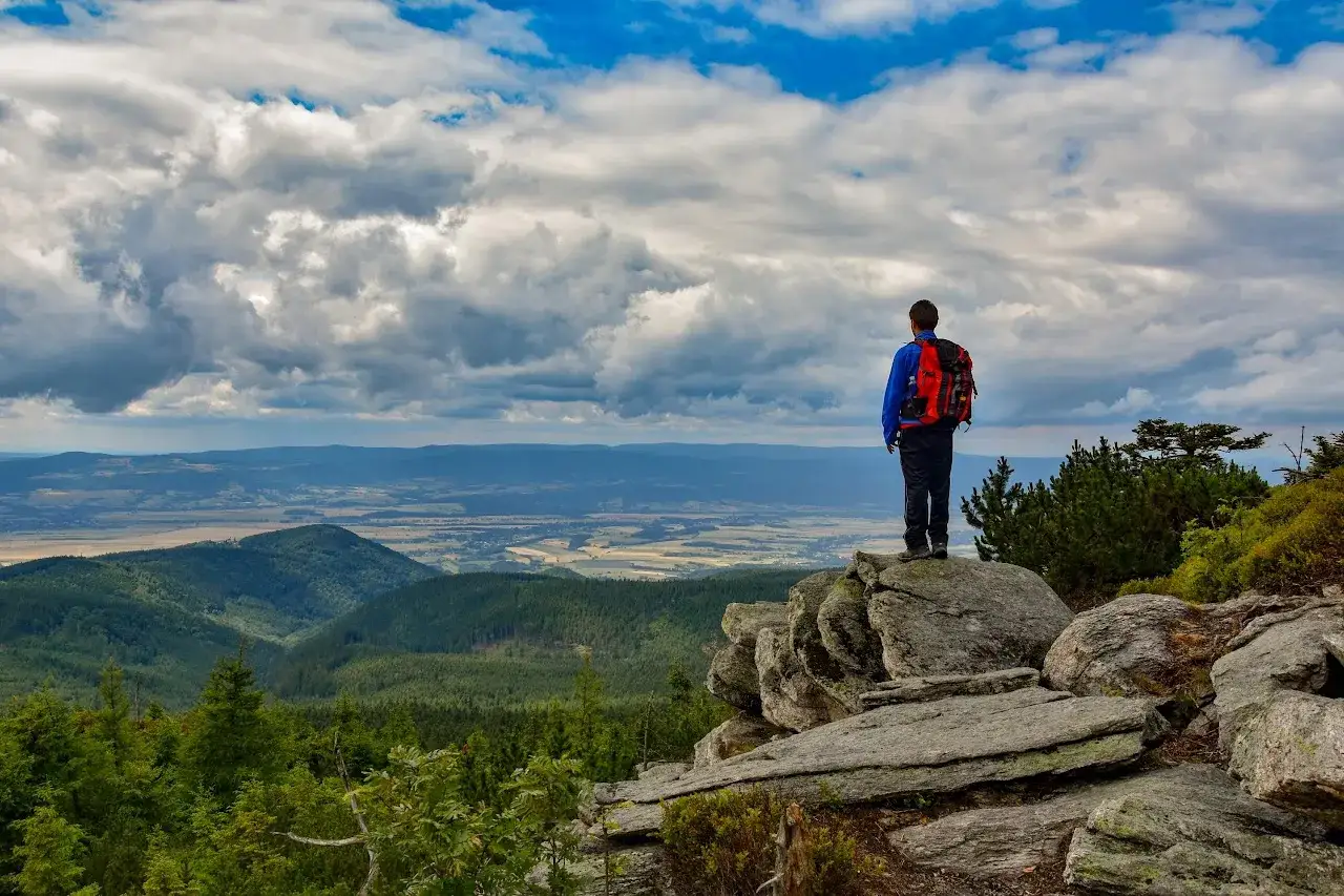 Turysta z plecakiem podziwia rozległą panoramę Sudetów, widząc malownicze miejscowości i lasy.