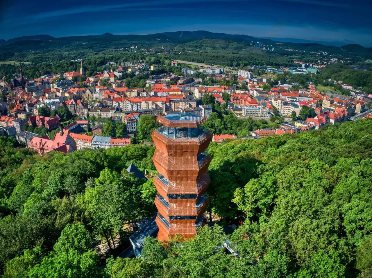 Nowoczesna wieża widokowa w otoczeniu zieleni, z panoramą miasta i Gór Wałbrzyskich w tle.