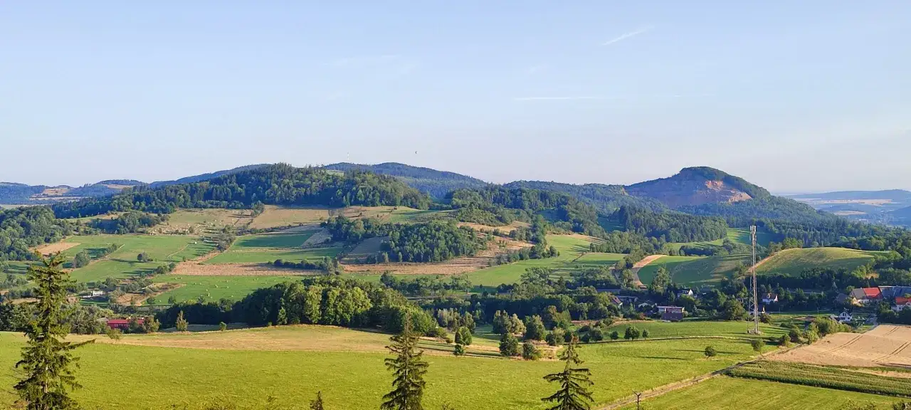 Zielone wzgórza i pola tworzą malowniczy krajobraz pogórza. W oddali widać lasy i niewielkie domy.