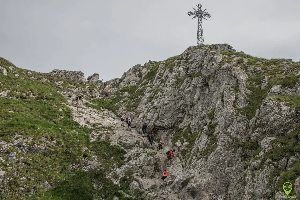 Grupa turystów wspina się po skalistym szlaku na Giewont. W oddali widać charakterystyczny krzyż.