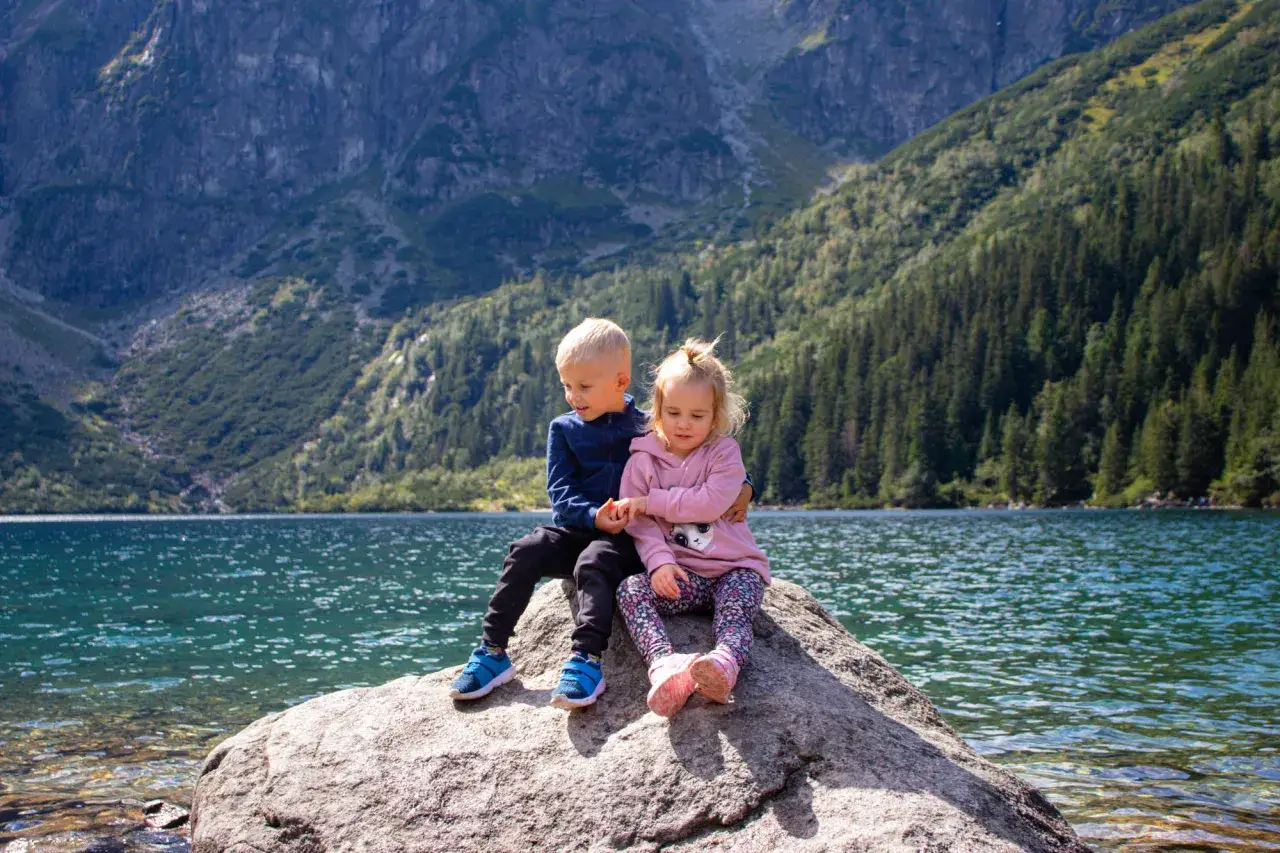 Dwoje dzieci siedzi na kamieniu nad Morskim Okiem, otoczeni górami i lasem.
