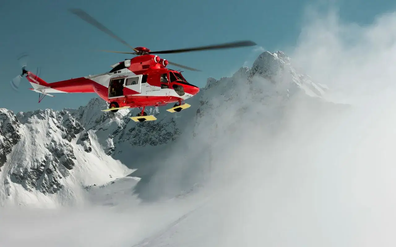 Czerwony helikopter ratunkowy w akcji, zapewniający pierwsza pomoc w górach. W tle ośnieżone szczyty i chmury.