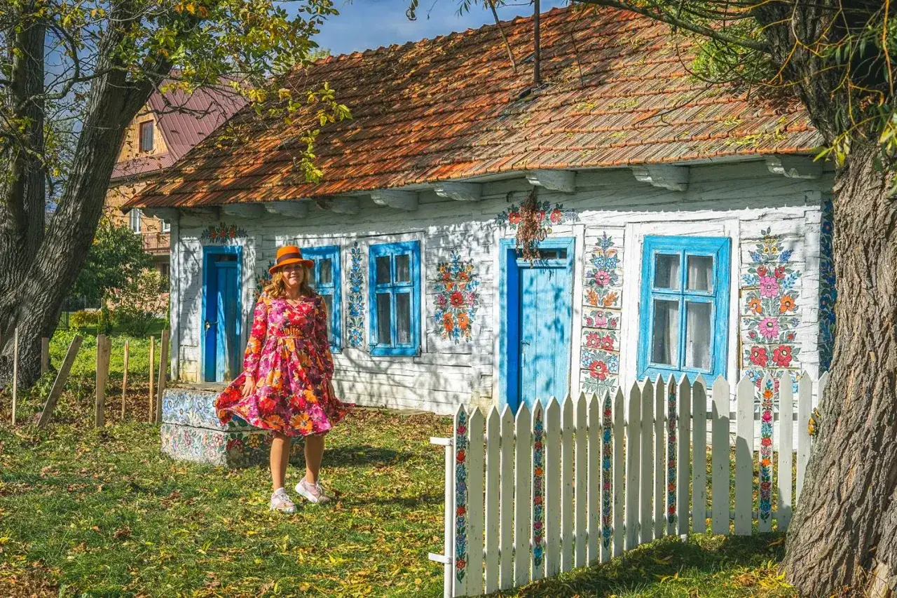 Kobieta w kolorowej sukience i kapeluszu podziwia malowane kwiaty na drewnianym domu podczas zwiedzania Zalipia.