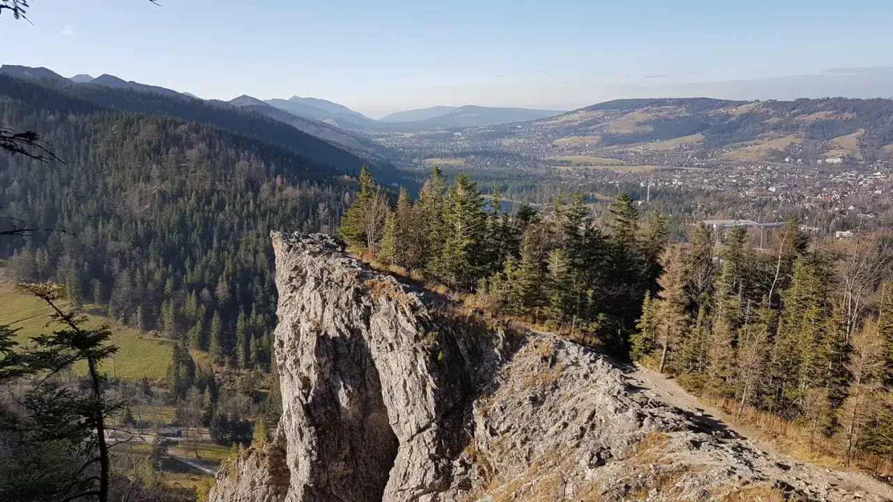 Widok z Nosala, góry samobójców, na Tatry i Zakopane. Skalisty szczyt, las iglasty i dolina z zabudowaniami.