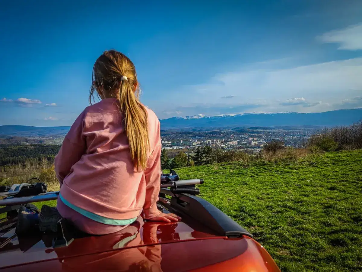 Dziewczynka siedzi na dachu samochodu, podziwiając panoramę miasta i gór. Idealny punkt widokowy Karpacz samochodem.